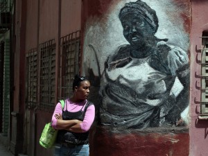 Una mujer permanece junto a un grafiti pintado en la pared de una vivienda, que muestra la imagen de una afrodescendiente, en La Habana Vieja, en la capital de Cuba. Crédito: Jorge Luis Baños/IPS