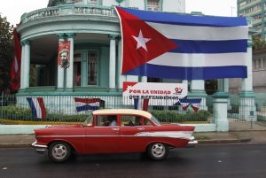 Un auto Chevrolet 1957, de fabricación estadounidense, utilizado como taxi colectivo transita próximo a la sede de la Dirección Nacional de Los Comités de Defensa de La Revolución Cubana (C.D.R); La imagen del líder de la revolución cubana Fidel Castro y una bandera cubana decoran la edificación situada en una céntrica calle del capitalino barrio del Vedado, en La Habana, Cuba el 27 de Noviembre de 2017. Crédito: Jorge Luis Baños/IPS.