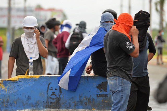 Jóvenes manifestantes nicaragüenses ocultan sus rostros detrás de una barricada en el sur de Managua, levantada con un muro en el que está impresa una imagen del presidente Daniel Ortega. La represión contra las protestas suma ya 131 personas muertas. Crédito: Jader Flores/IPS