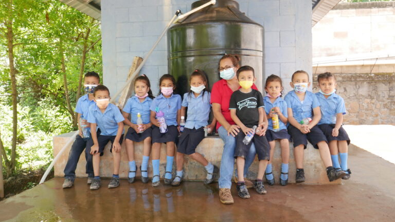 La maestra Angélica María Posada, directora de la escuela del caserío El Guarumal, ubicado en el municipio de Sensembra, en el departamento de Morazán, en el este de El Salvador, posa junto a algunos de sus alumnos de primaria delante del tanque que abastece de agua potable a ese centro de estudios y también a 150 familias de ese y otros caseríos aledaños. El agua lluvia es captada en el techo de lámina y dirigida a una cisterna subterránea, luego es filtrada en un cuarto especial y bombeada al tanque con grifo, lista para el consumo. Foto: Edgardo Ayala / IPS